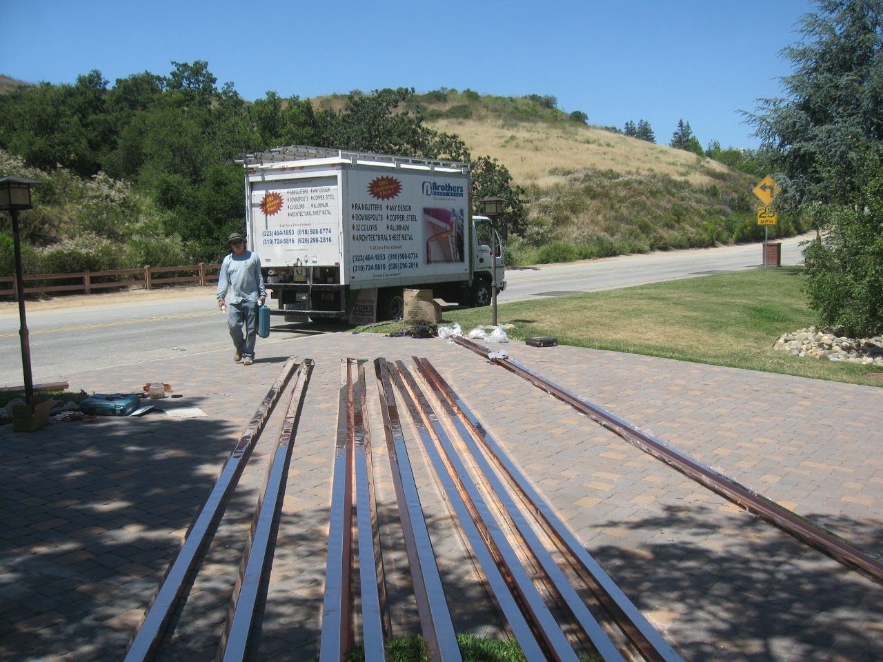 Man walks toward a truck on a gravel road with long metal pieces laid out; a grassy hill in the background.