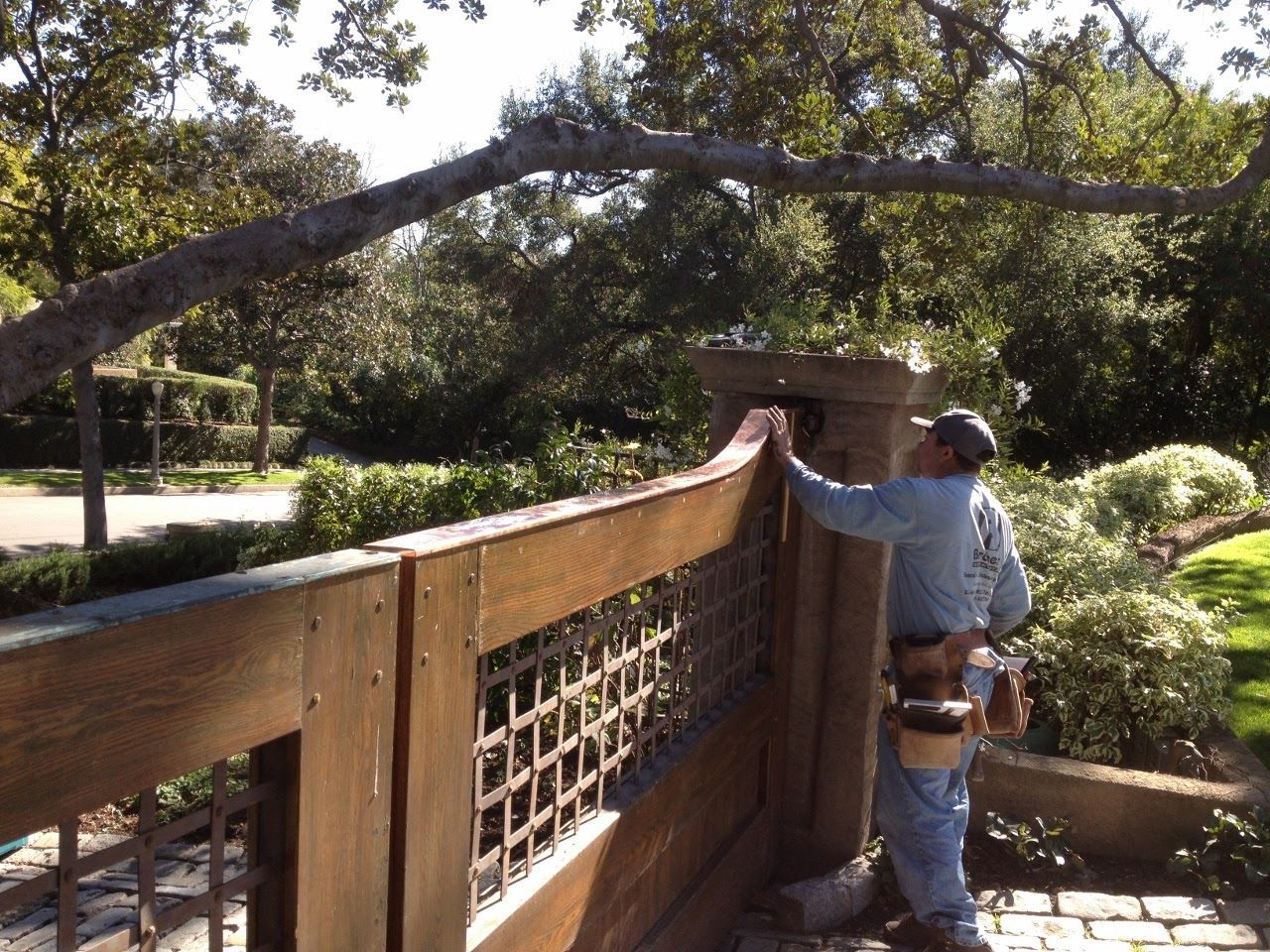 Man working on a wooden fence, outdoors.  Green trees and bushes in background. Sunny, daytime.