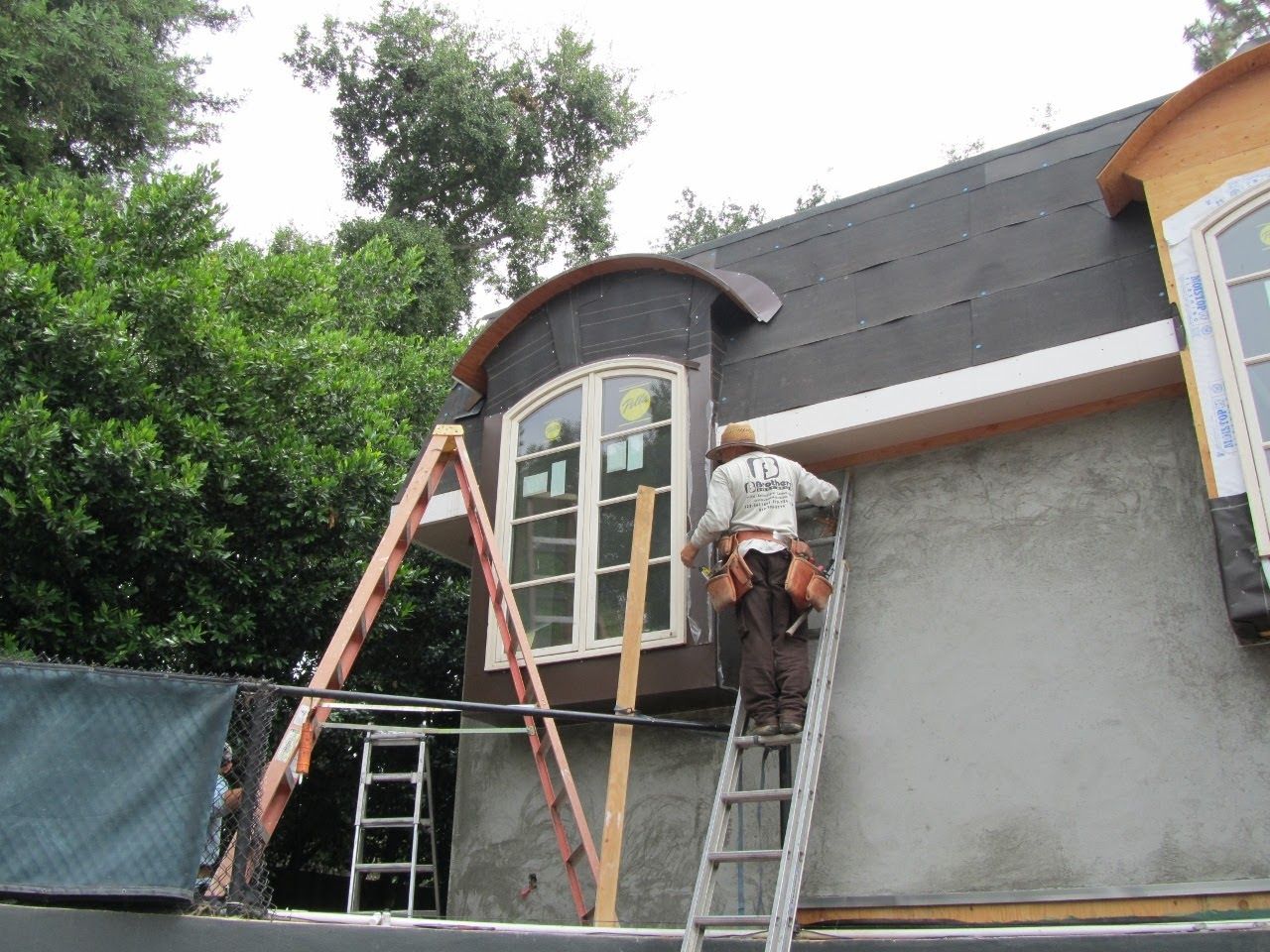 Construction worker on ladder, installing roofing on house exterior; brown, grey, and white.
