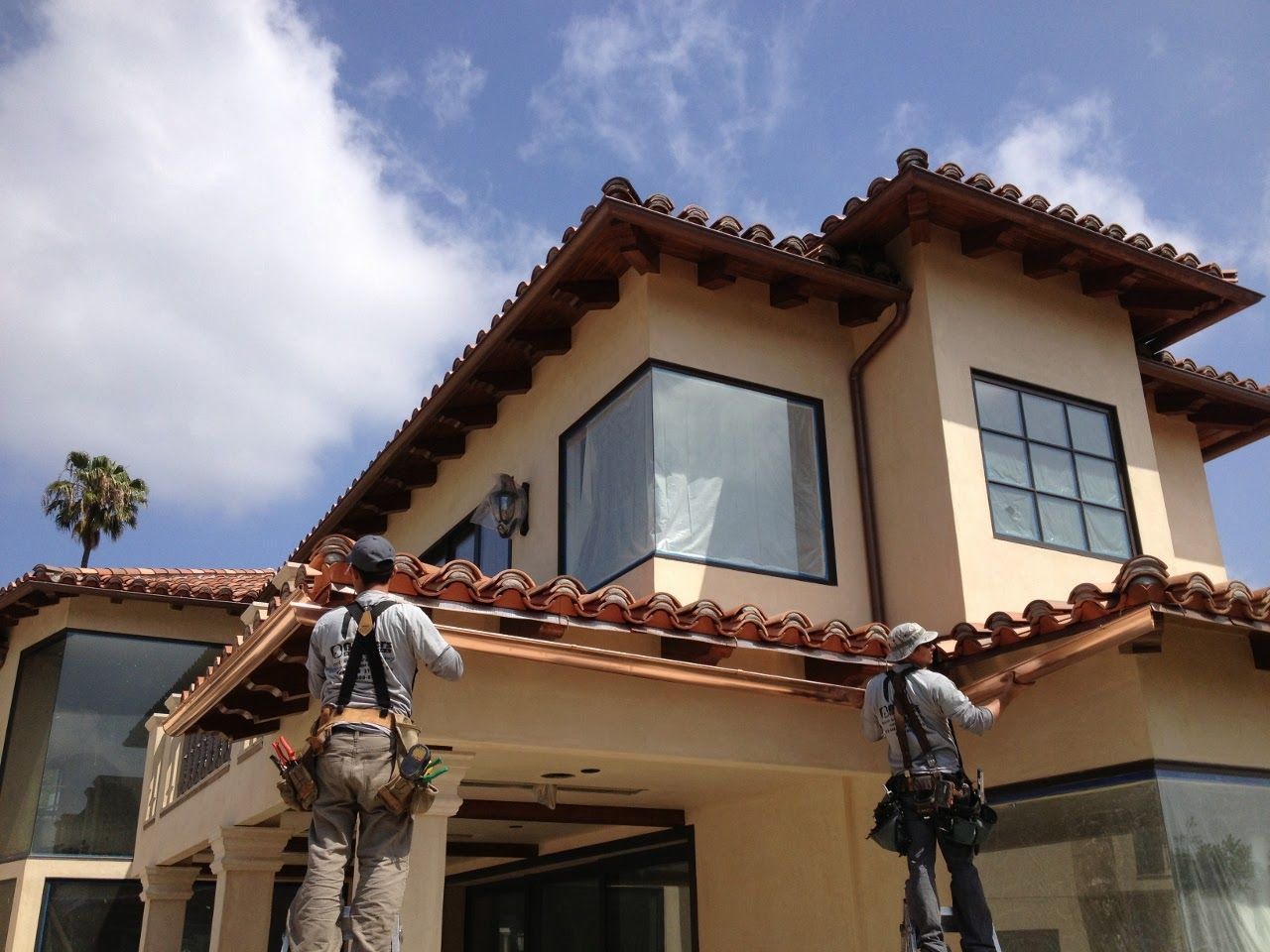 Two workers installing gutters on a two-story Spanish-style house under a blue sky.