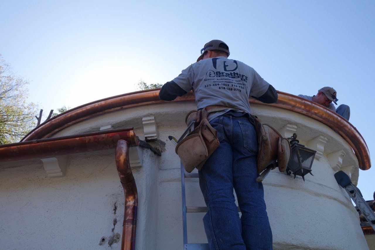 Two workers install copper roofing on a white, curved building with matching gutters.