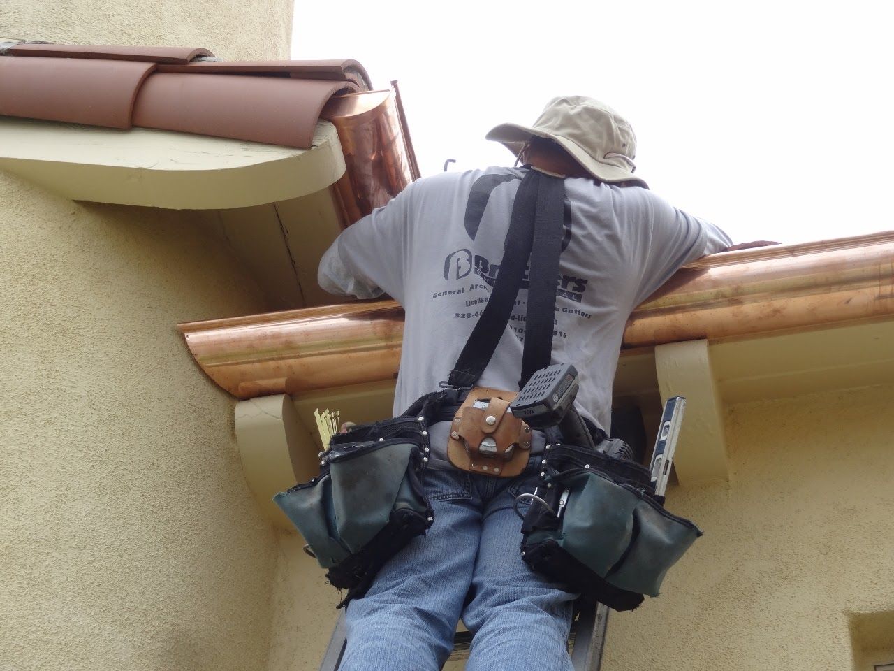 Man on a ladder installing copper gutters on a beige stucco building, wearing tool belt and tan hat.