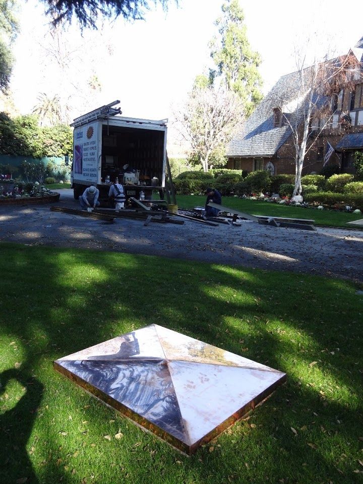 A square metal object on grass; moving van with workers in the background near a house.