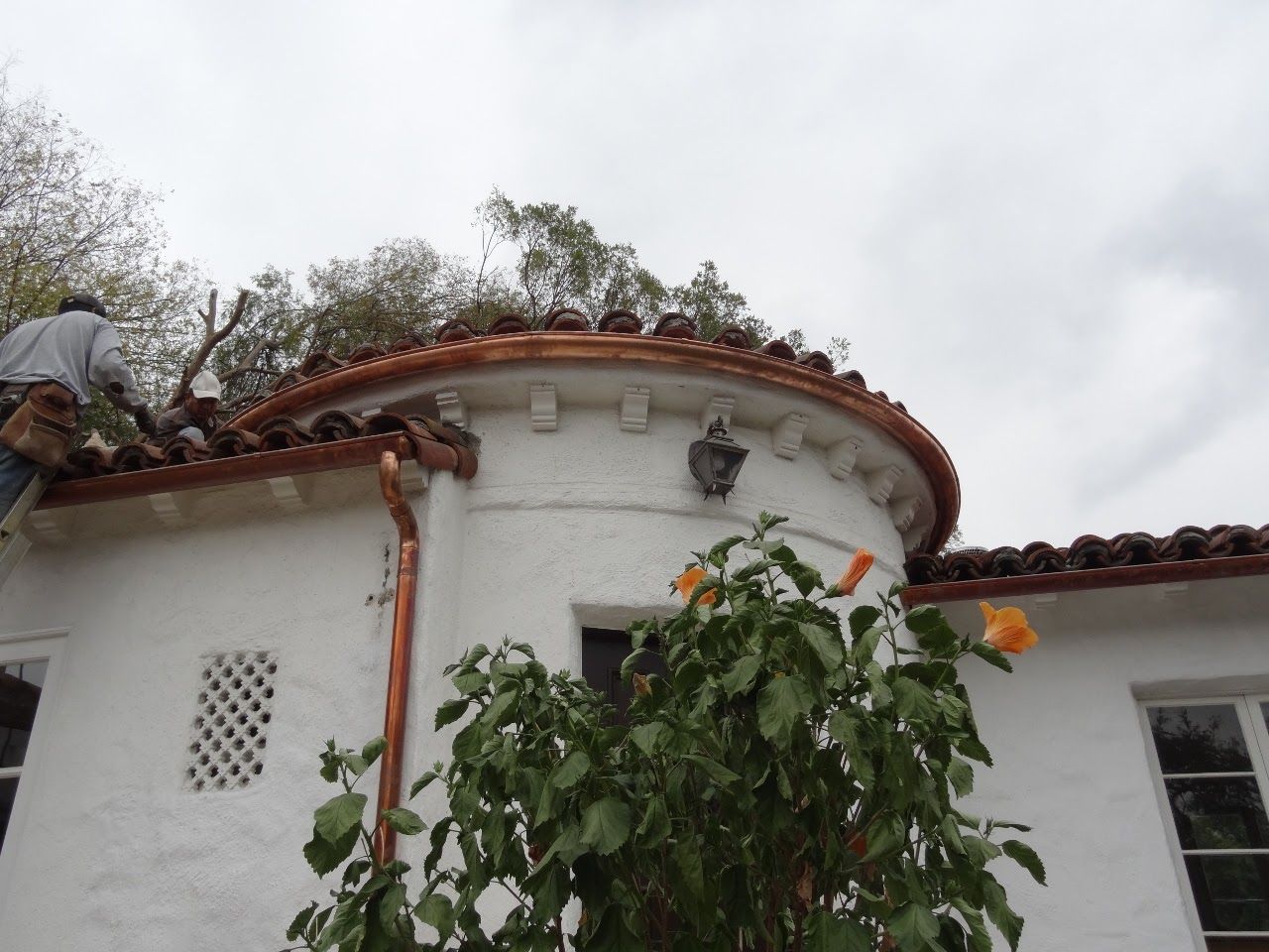 Person on roof of white stucco building with copper gutters, tiles, and turret with trees in background.