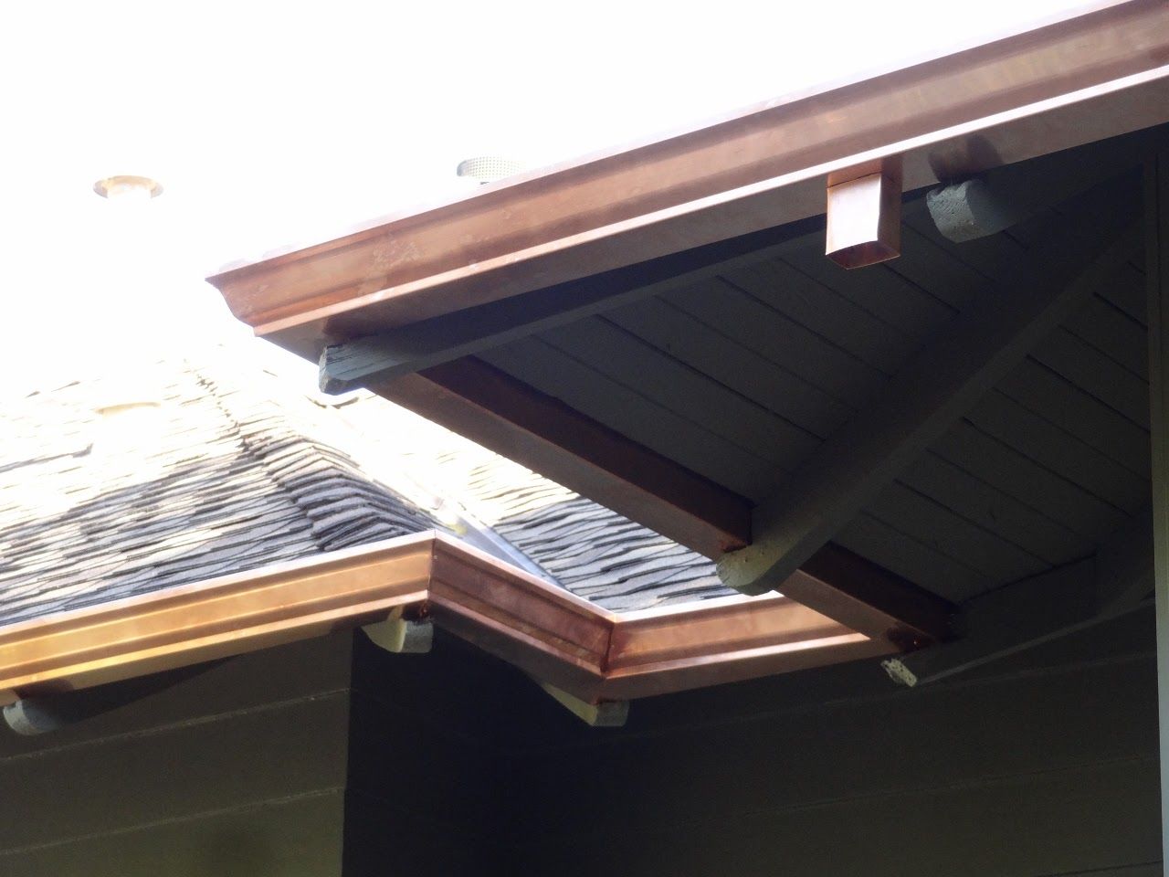 Copper gutters on a roof's edge, angled towards a corner. Sunlight catches the metal.