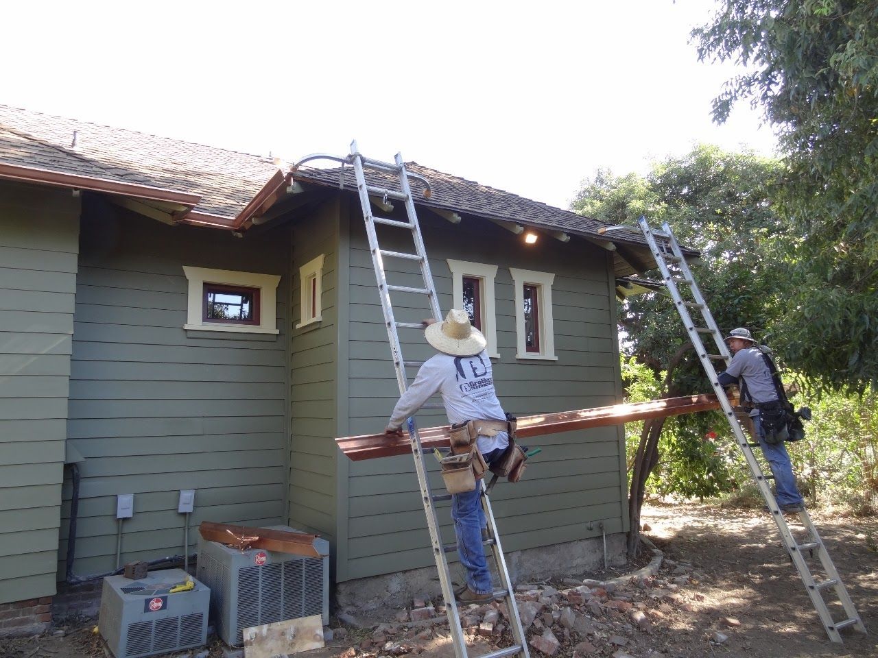 Two construction workers on ladders installing a wooden beam on a green house exterior.