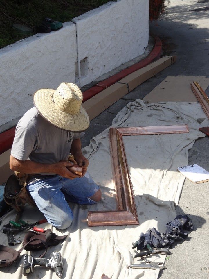 Man on knees assembling a copper frame outdoors, wearing a straw hat. Tools and materials scattered.
