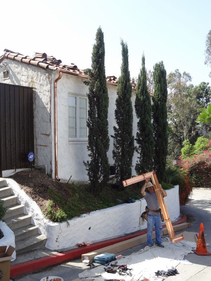 Man holding a copper frame, standing near a stucco house with cypress trees; tools and materials are on the ground.