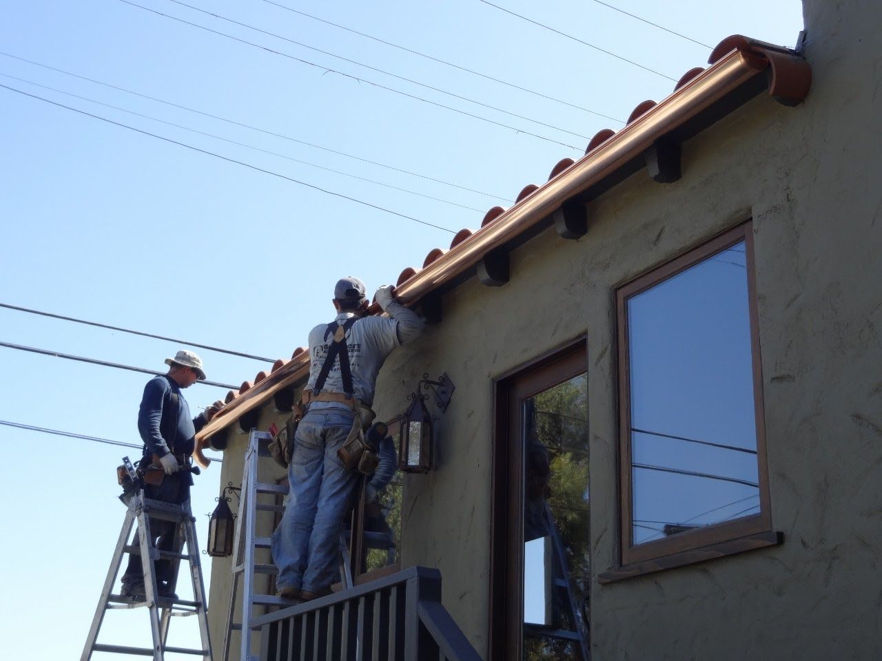 Two workers installing copper gutters on a stucco house near power lines under a blue sky.