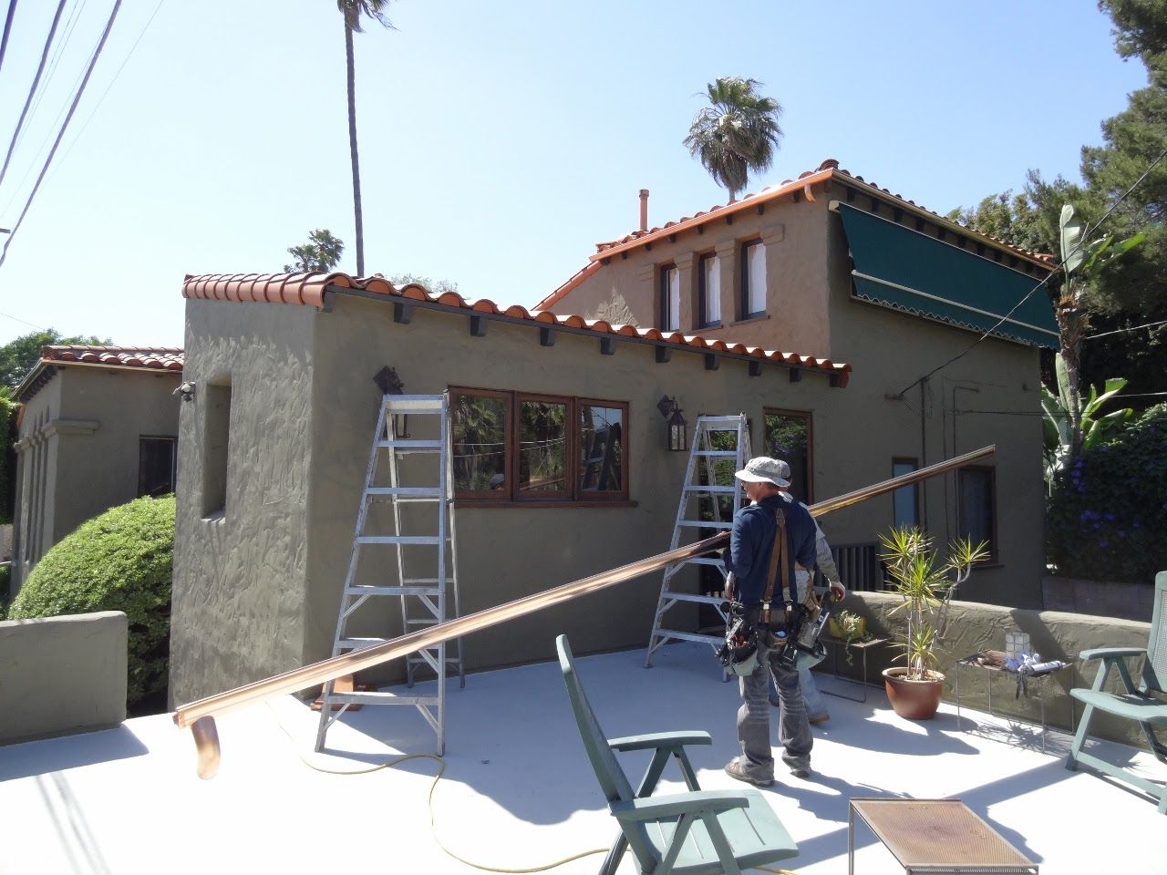 Man working on a roof with ladders, brown wood beam, and a green stucco building.