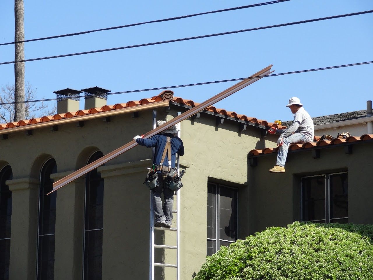 Construction workers installing metal flashing on a roof, one on a ladder, other sitting on the edge.