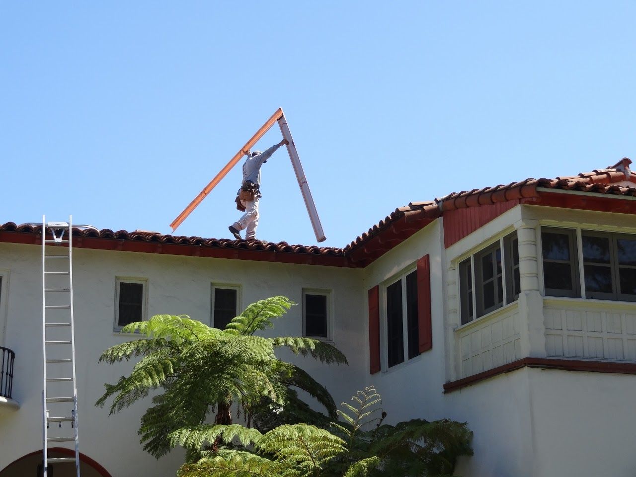 A person on a roof balancing a long piece of lumber in the sun. A ladder sits nearby.