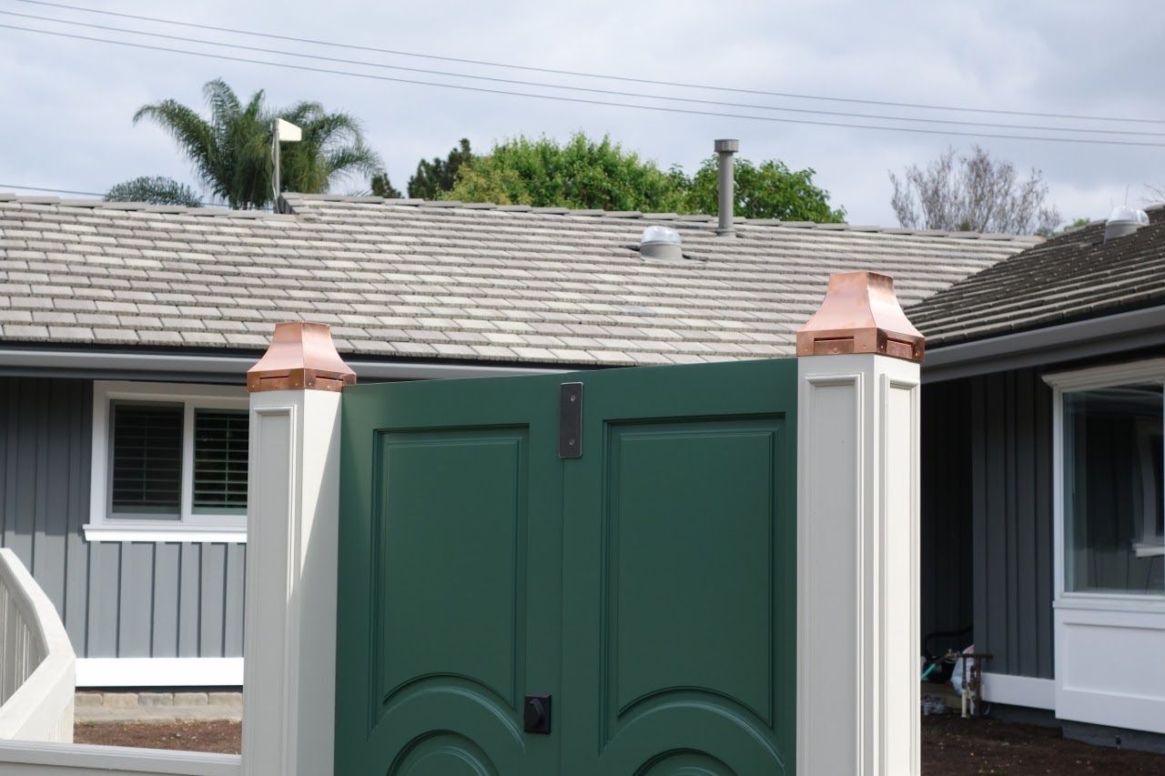Green gate with white pillars, topped with copper caps, in front of a gray house.