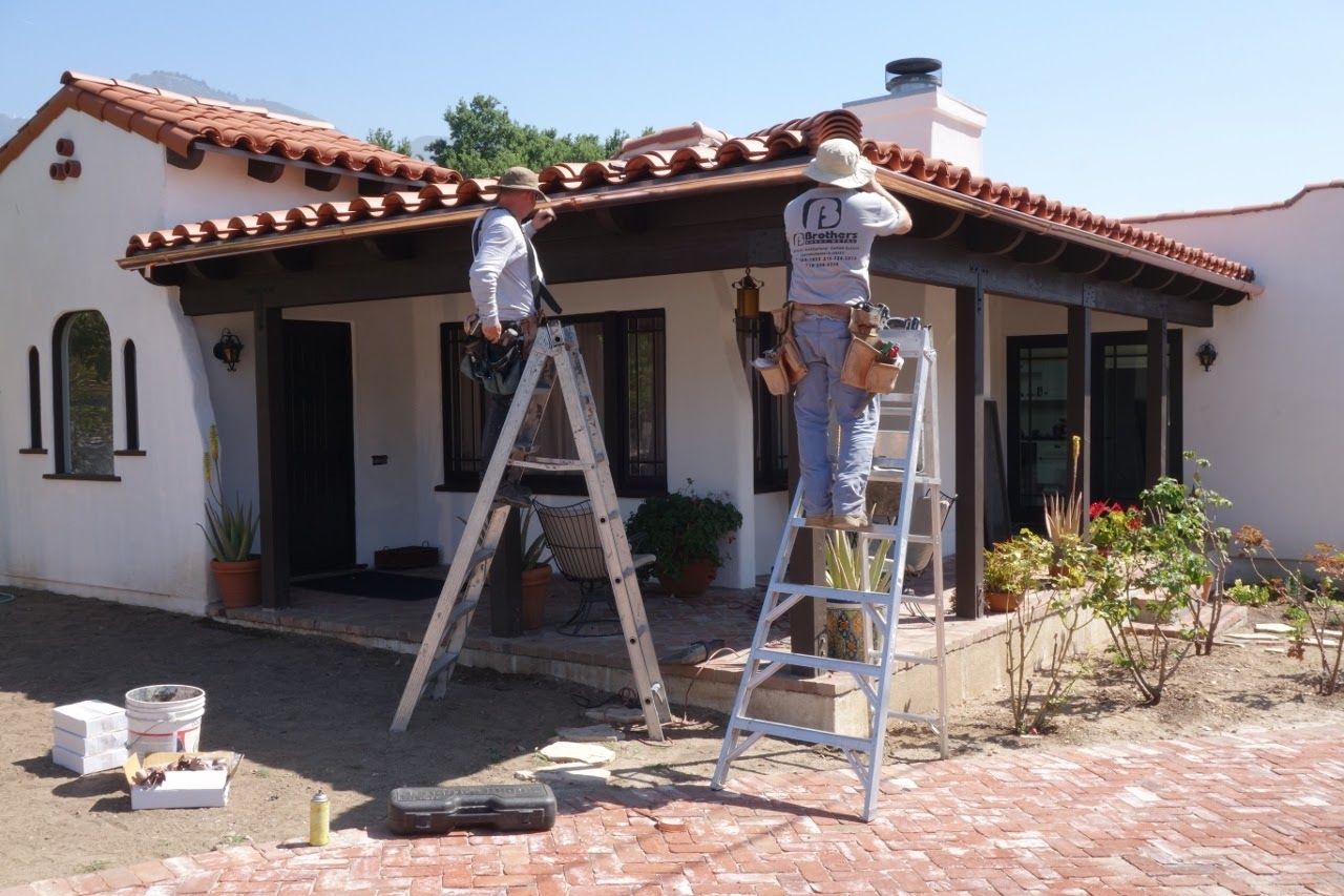 Two workers on ladders repairing a Spanish-style home's roof. Brown tiles, white stucco, and a sunny day.