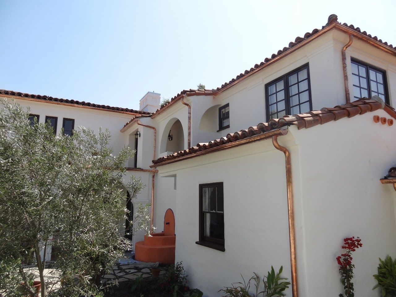 White stucco house with copper gutters, red tile roof, black windows, and olive tree.