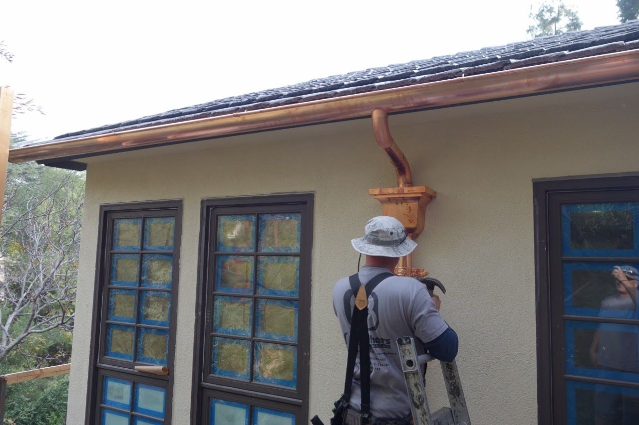 Man on ladder installing copper gutter on a stucco building with blue-framed windows.