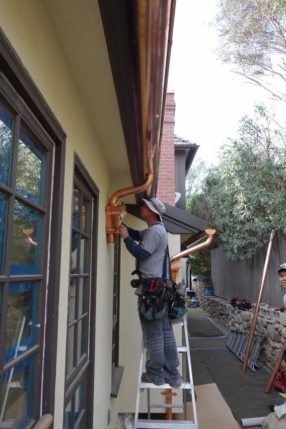 Man on ladder installs copper gutter near window and tree.