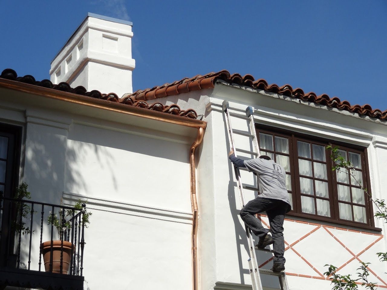 Person on ladder, inspecting the roofline of a white stucco building with a copper gutter and red tile roof.