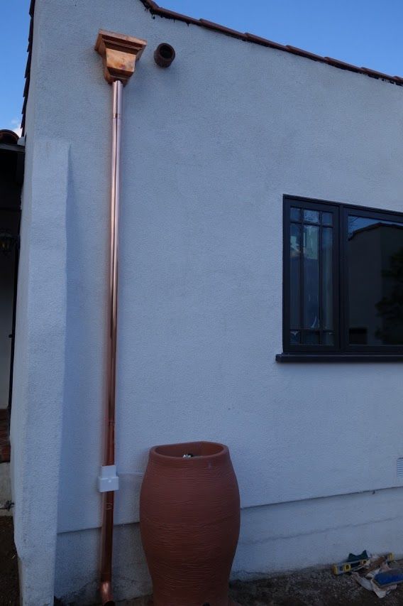 Copper rain gutter and downspout on a white stucco house, with a terracotta pot below.