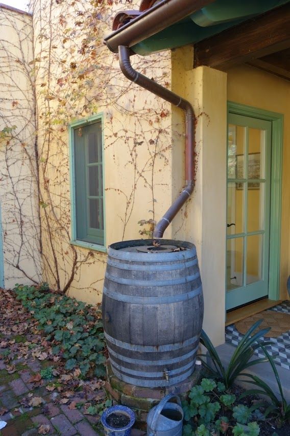 Rain barrel collecting water from a copper gutter, next to a yellow building with a green door.