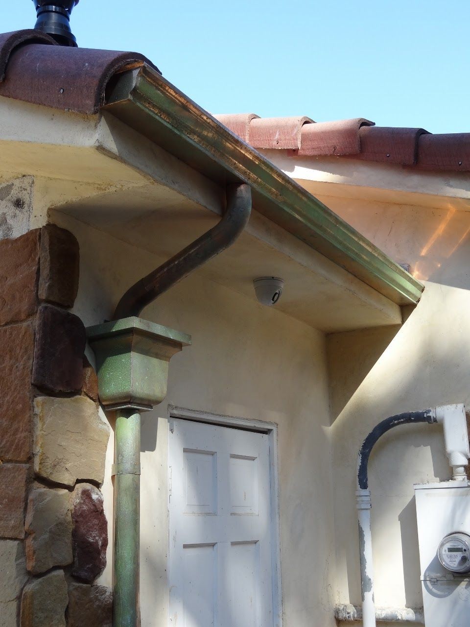 Green-patinaed copper gutters and downspout on a stucco building with a white door and stone pillar.