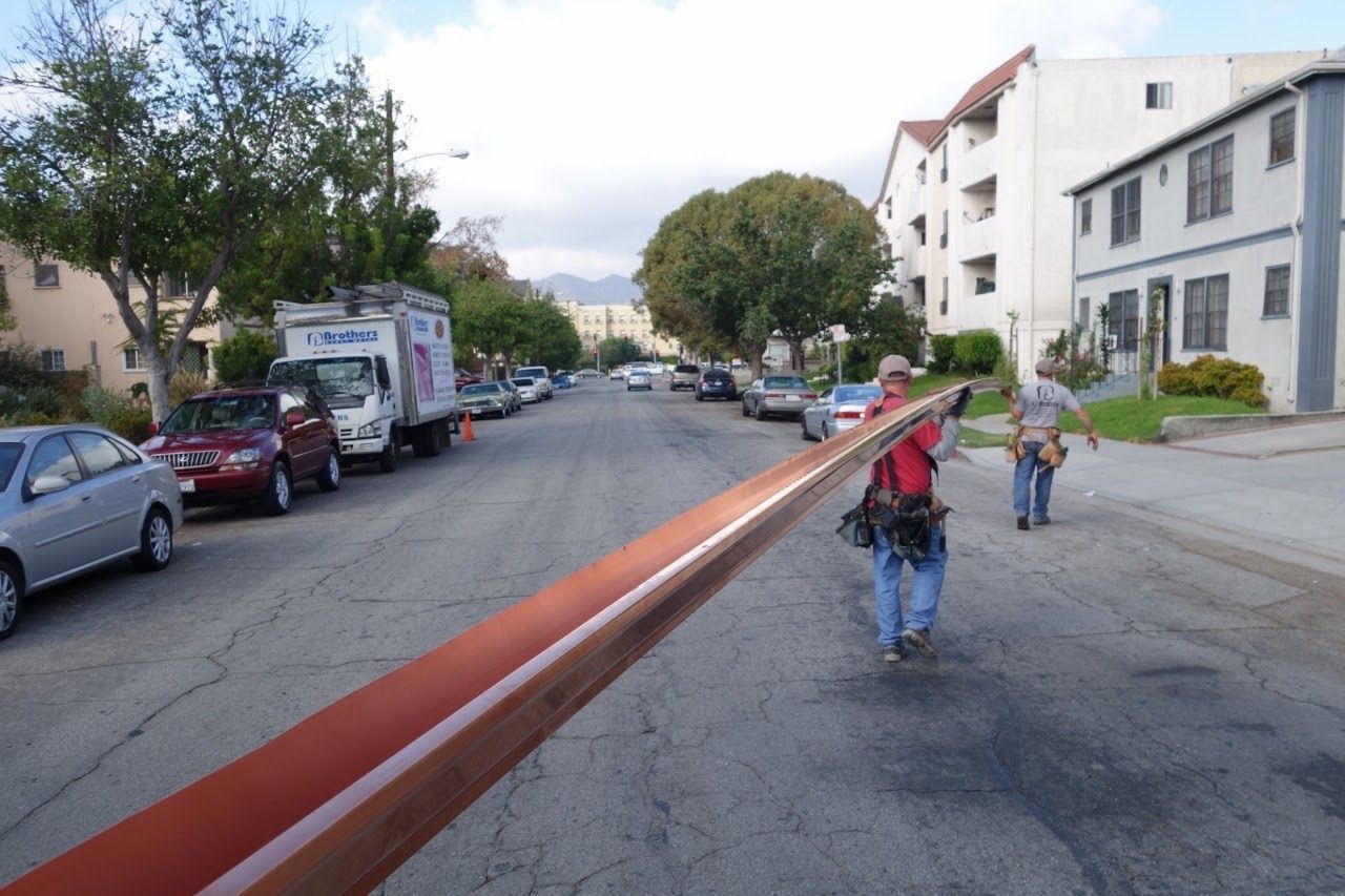 Three people carrying a long, copper-colored beam down a street lined with parked cars and buildings.
