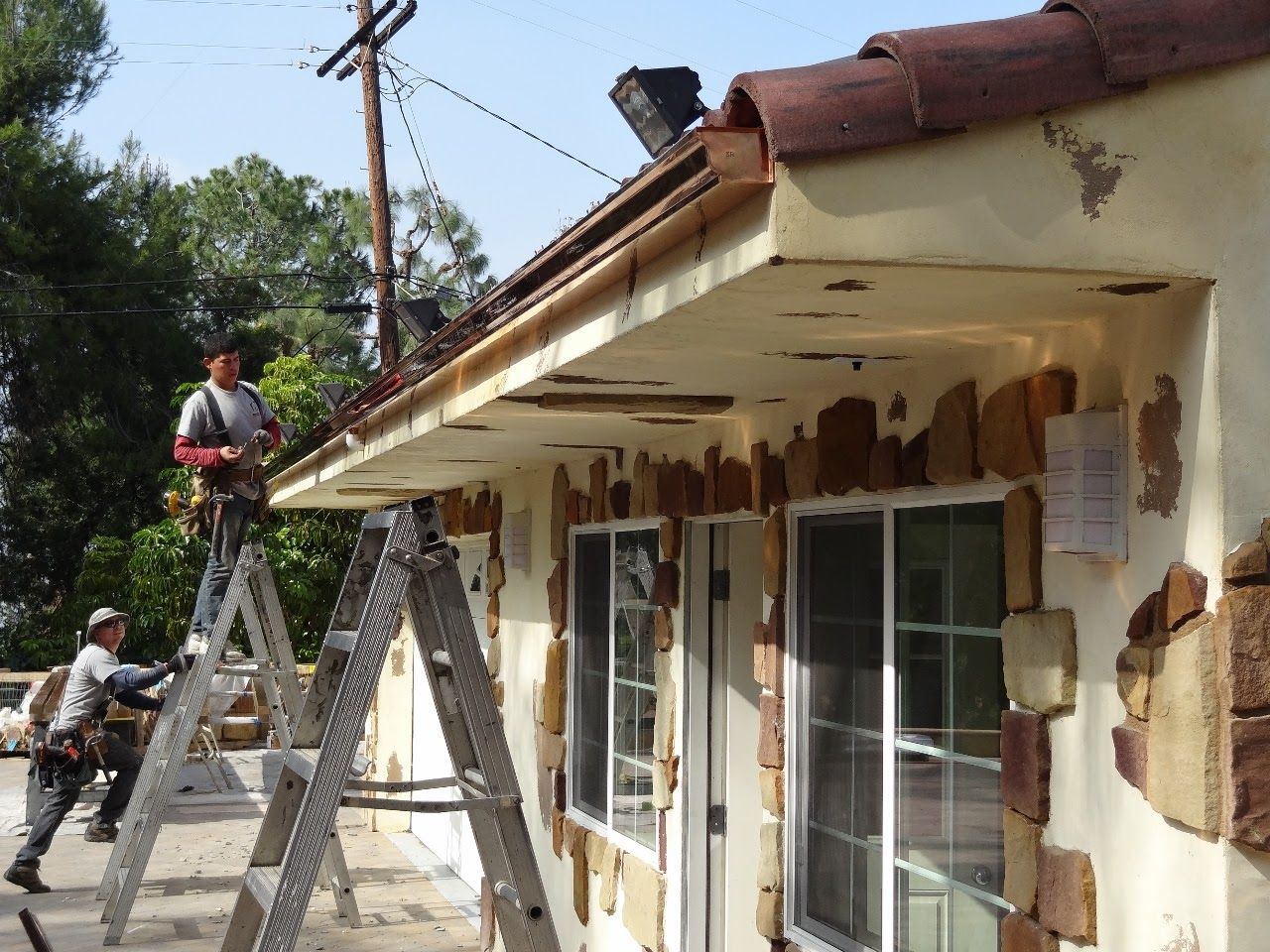 Two workers on ladders repairing the roofline of a stucco building.