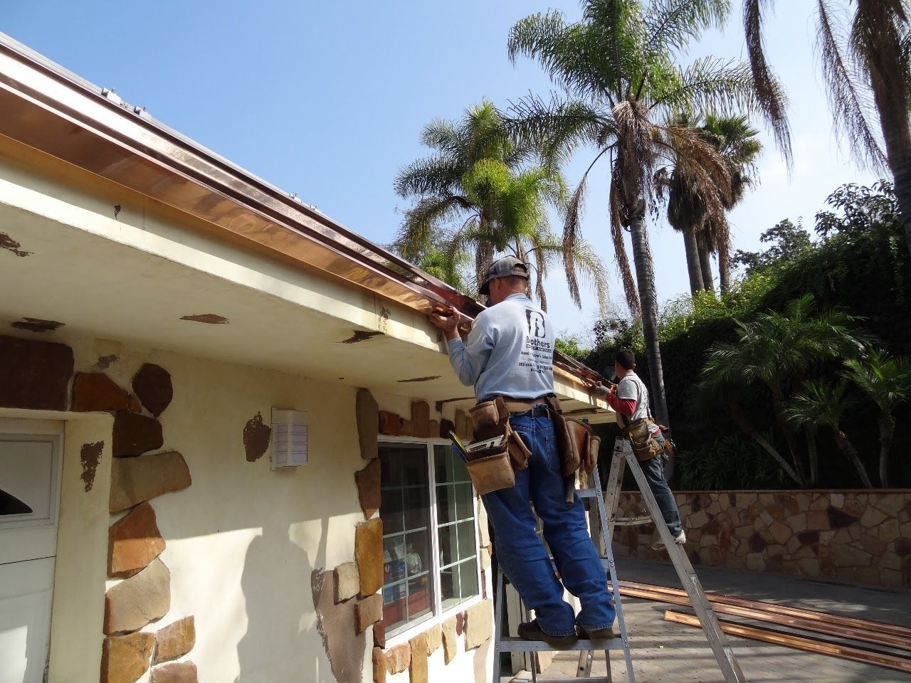 Two workers installing copper gutters on a home. One on ladder, other on the roof, sunny outdoors.