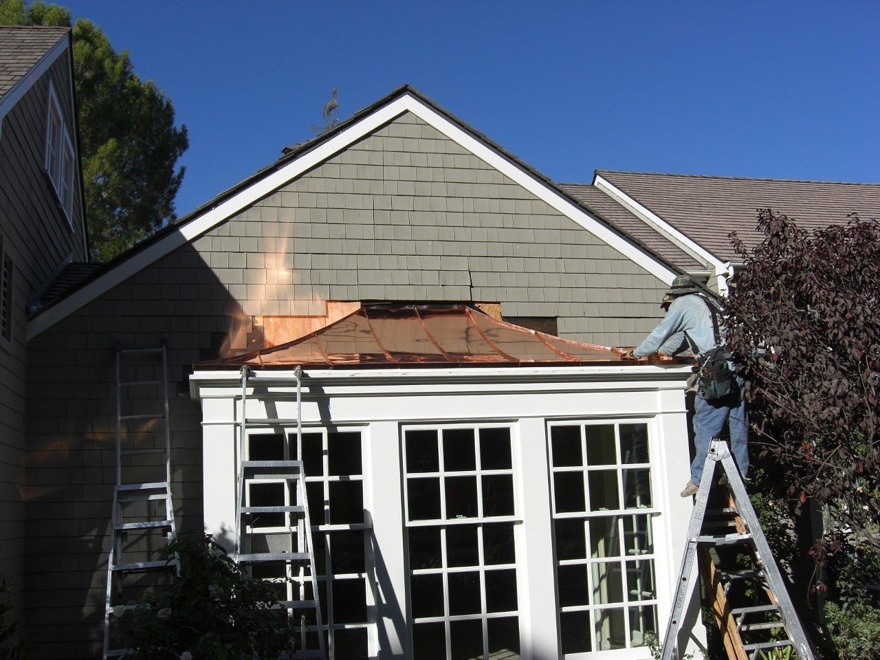 Roofer on a ladder working on a copper roof, with white-framed glass doors below; blue sky.