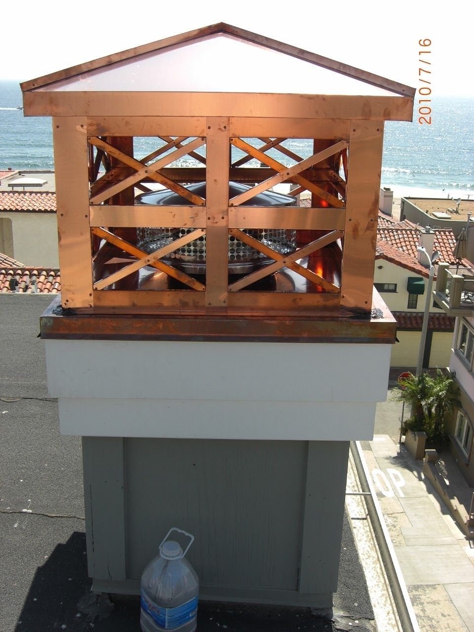 Copper chimney cap on a white and gray structure, with ocean view in background.