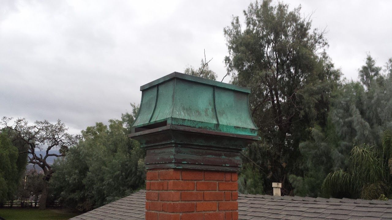 Brick chimney with green copper cap against a cloudy sky and tree backdrop.