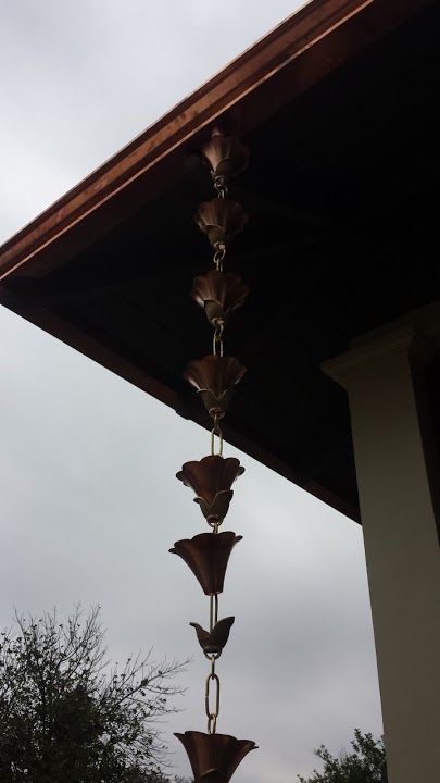 Copper rain chain with flower-shaped cups hanging from a brown gutter against a cloudy sky.