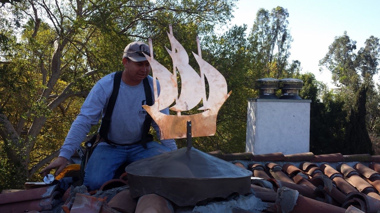 Man on a roof installing a copper weathervane shaped like a ship. Sunny day, trees in background.