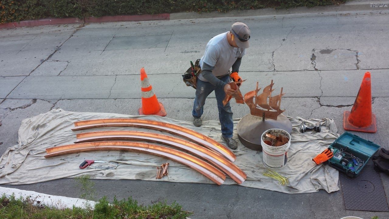 Man working on copper pieces outdoors, orange cones and tools present.