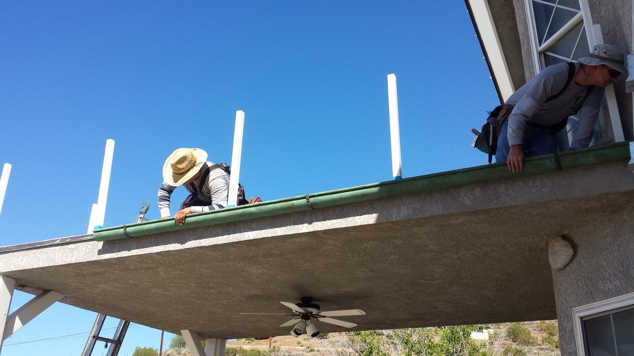 Two workers on a roof installing white posts; clear blue sky.