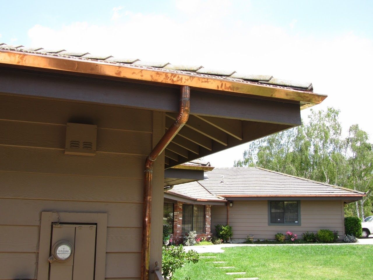 Brown house with rusty gutters and tiled roof. Green grass and trees in the background.