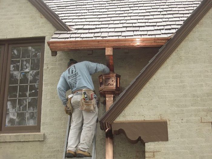 Man on ladder installing copper gutter near a window on a brick building.