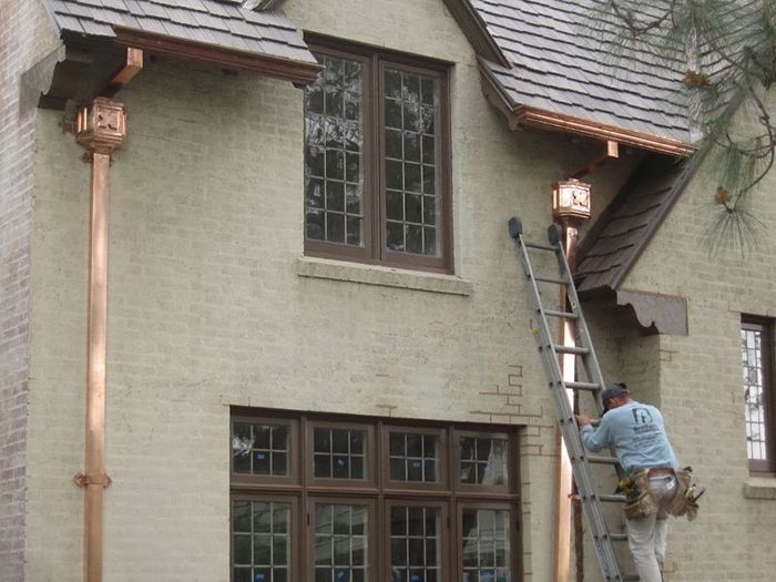 Man on ladder installing copper guttering on a two-story stucco house with brown trim.