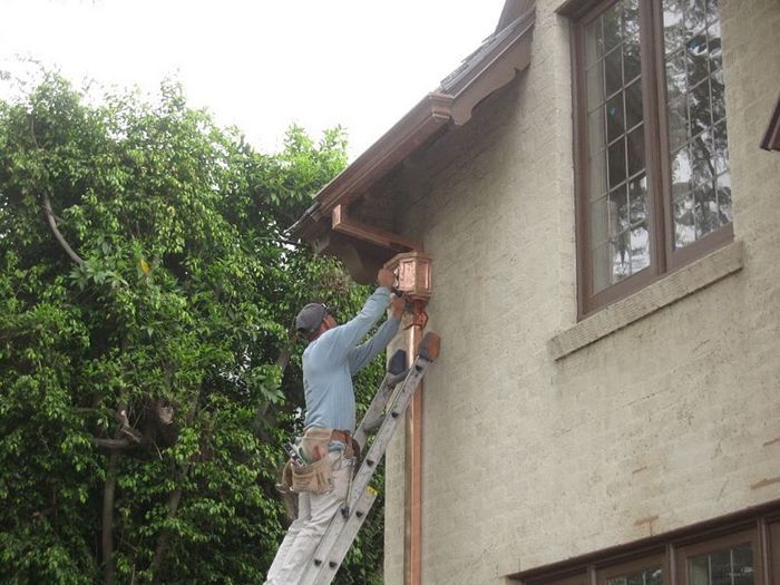 Man on a ladder installing a copper downspout on a stucco house.