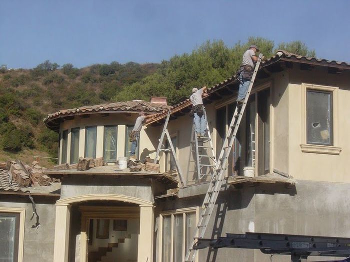 Three workers on ladders repairing roof tiles on a two-story beige stucco house with a hillside background.