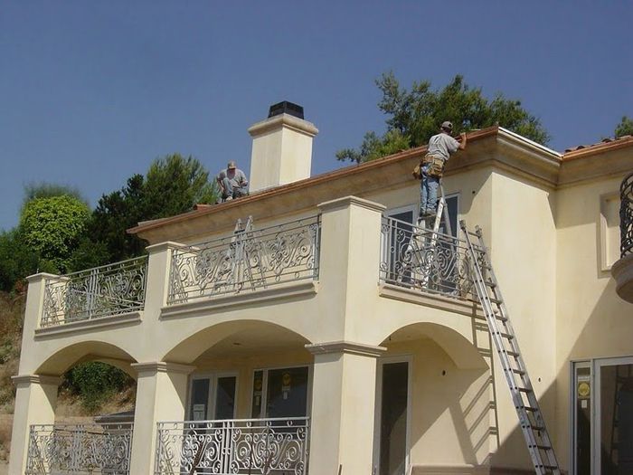 Two workers on a light yellow house's roof and balcony, one on a ladder. Blue sky, trees in the background.