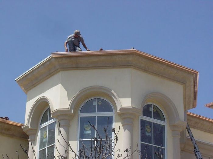 Man working on the roof of a beige house with arched windows, ladder visible. Sunny day.