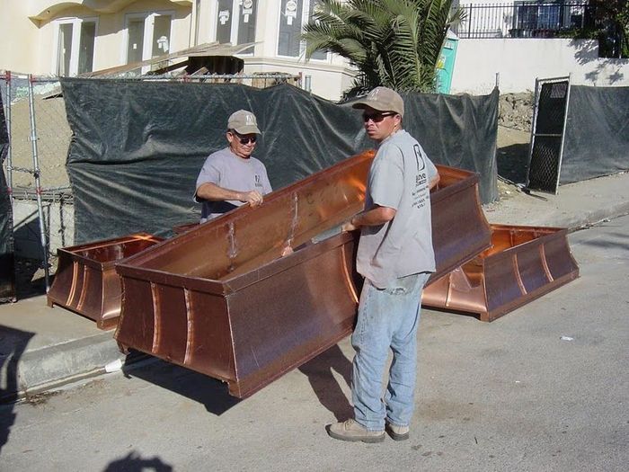 Two men carrying a large copper container outdoors near a construction site.