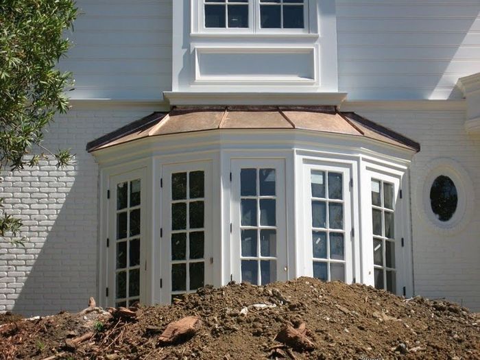 White house with bay window and copper roof, sitting in front of dirt.