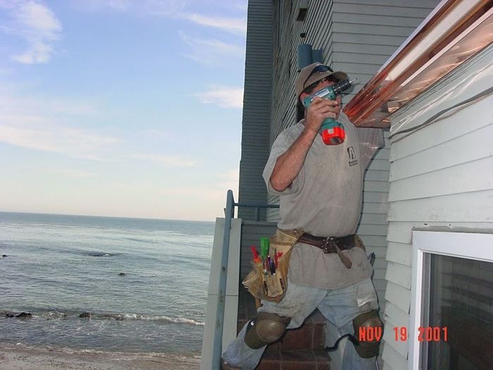 Man installing copper gutters on a house overlooking the ocean; he's using a power drill.