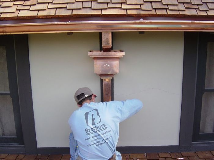 Man installing copper gutter on a beige building with dark window frames.