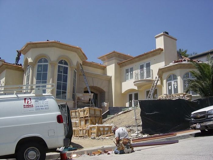 Large, stucco house under construction with workers, ladders, and construction materials on a sunny day.