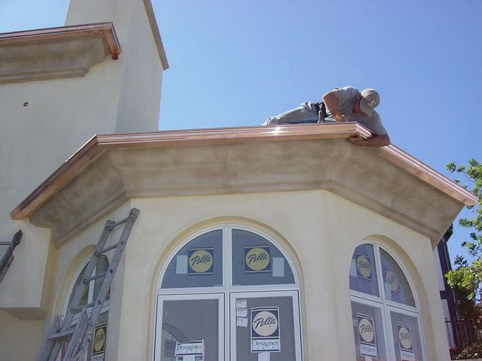 Man working on a copper-trimmed roof of a building with arched windows, under a bright blue sky.