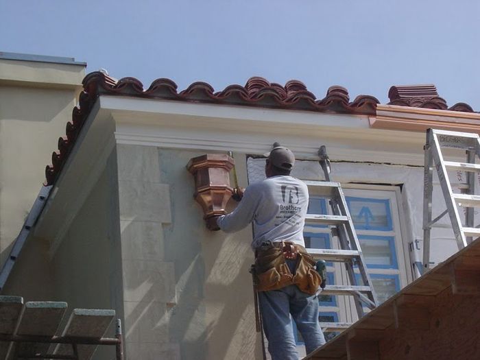 A construction worker on a ladder installing a copper light fixture on a stucco wall with a tiled roof.