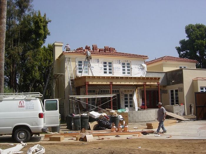House under construction, workers on roof and scaffolding. White van and trees in the background.